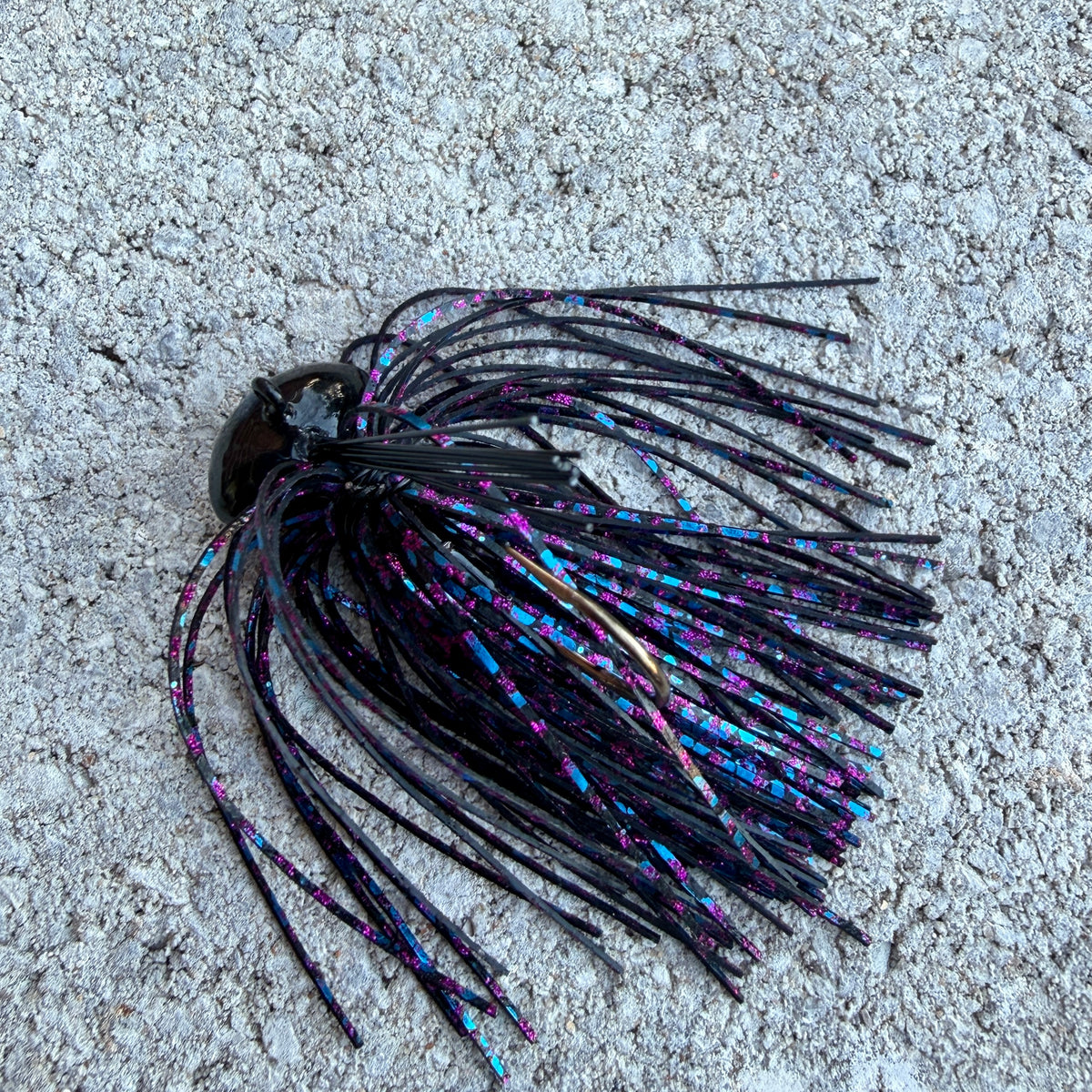 A single fishing lure with a black head and long black skirt strands tipped in iridescent blue, purple, and pink, photographed on a rough light gray concrete surface.