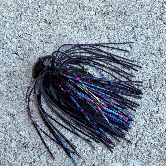 A single fishing lure with a black head and long black skirt strands tipped in iridescent blue, purple, and pink, photographed on a rough light gray concrete surface.