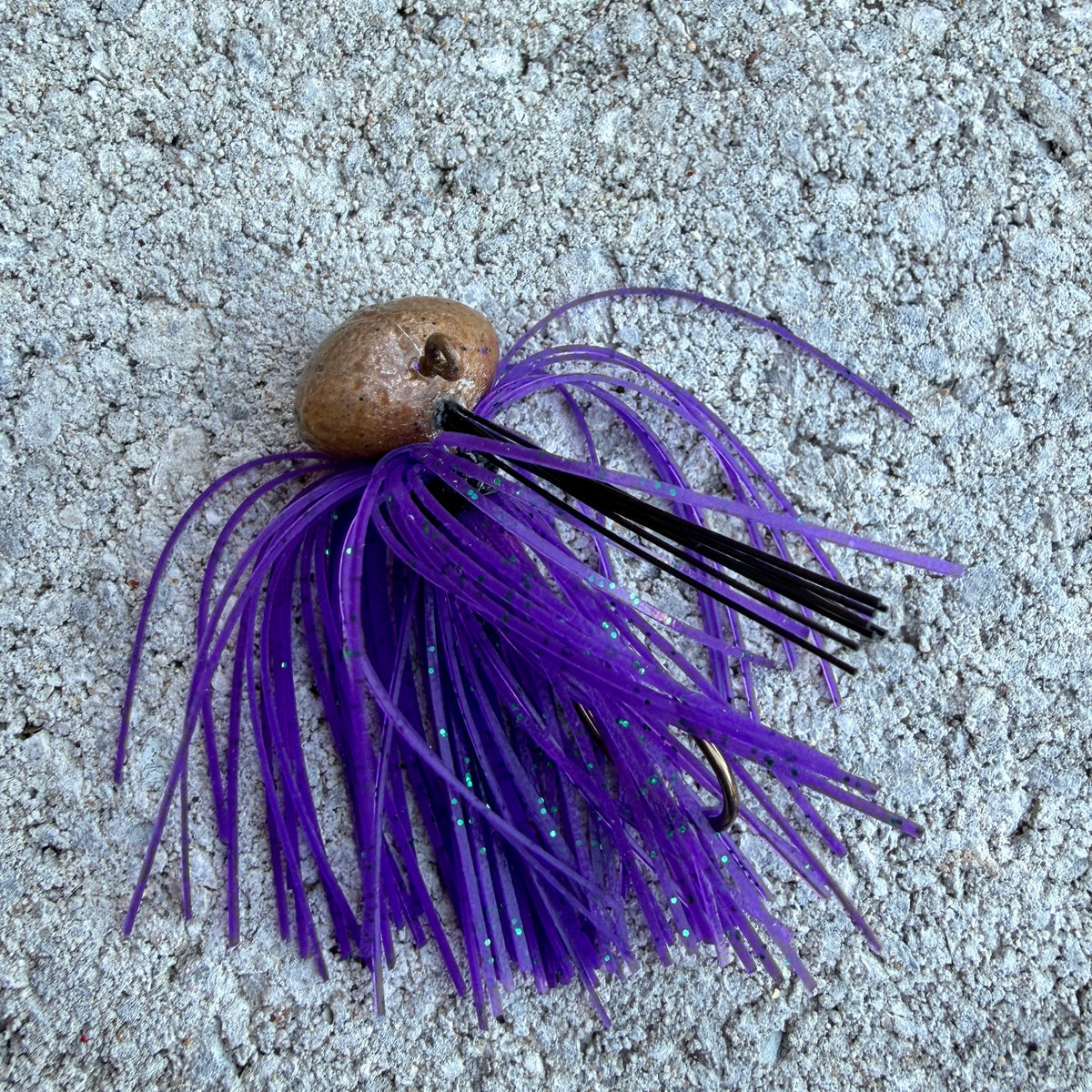 A fishing lure with a brown plastic body, black and purple skirt strands, and a visible hook, placed on a textured gray concrete surface.