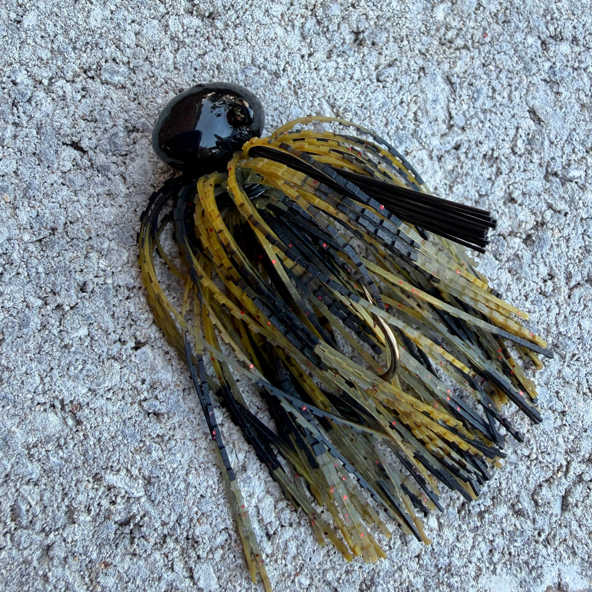 A close-up of a fishing jig lure with a glossy black head and a skirt made of black, olive green, and tan strands, lying on a textured gray concrete surface.