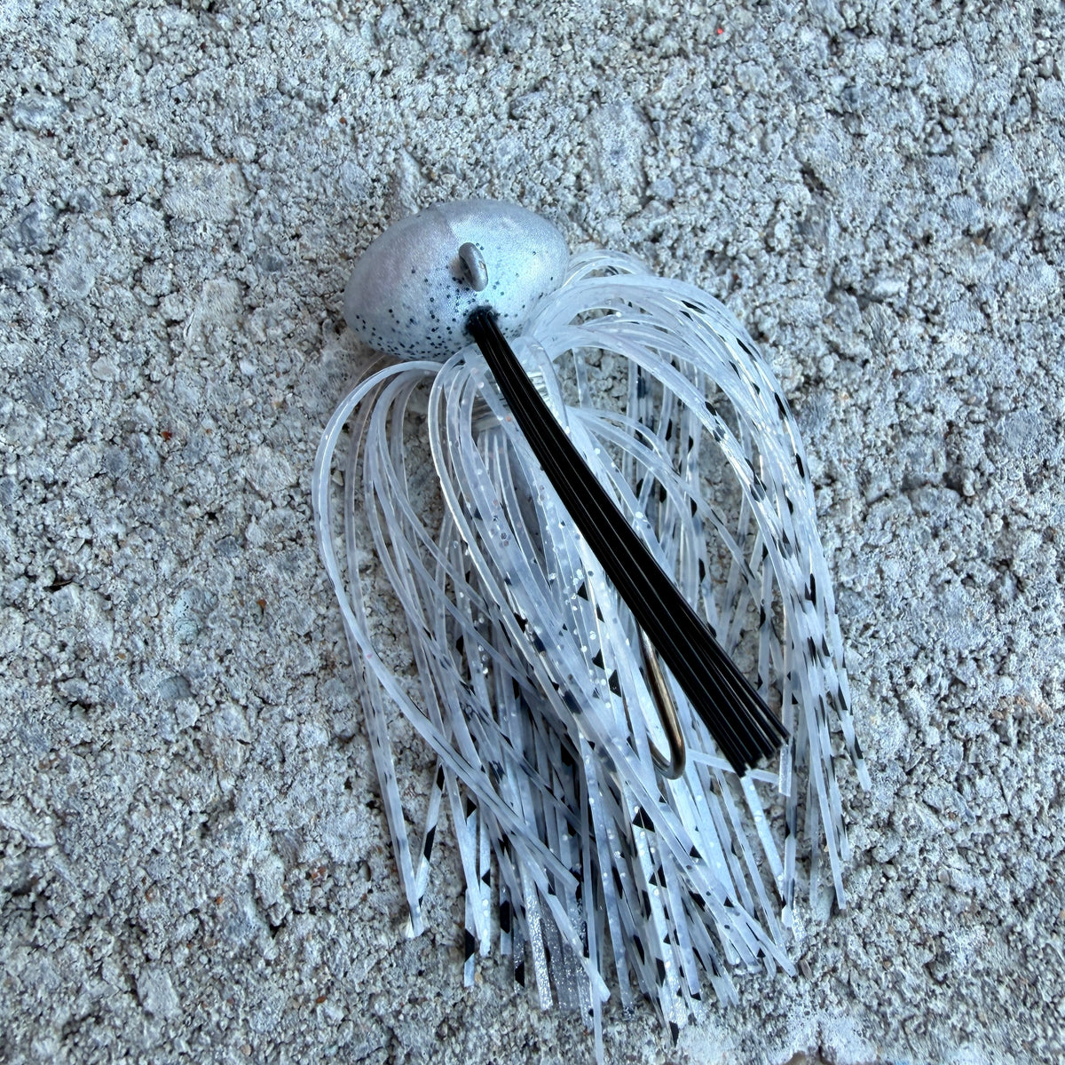 A fishing lure with a silver metal top, black center rib, and translucent white skirt with black speckles, photographed on a textured gray concrete surface.