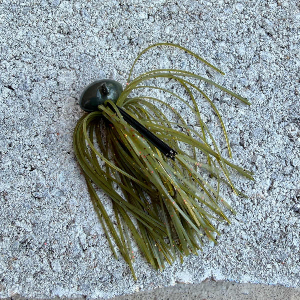 A soft plastic fishing lure with olive green skirt strands and a dark green to black top, photographed on a textured light gray concrete surface.