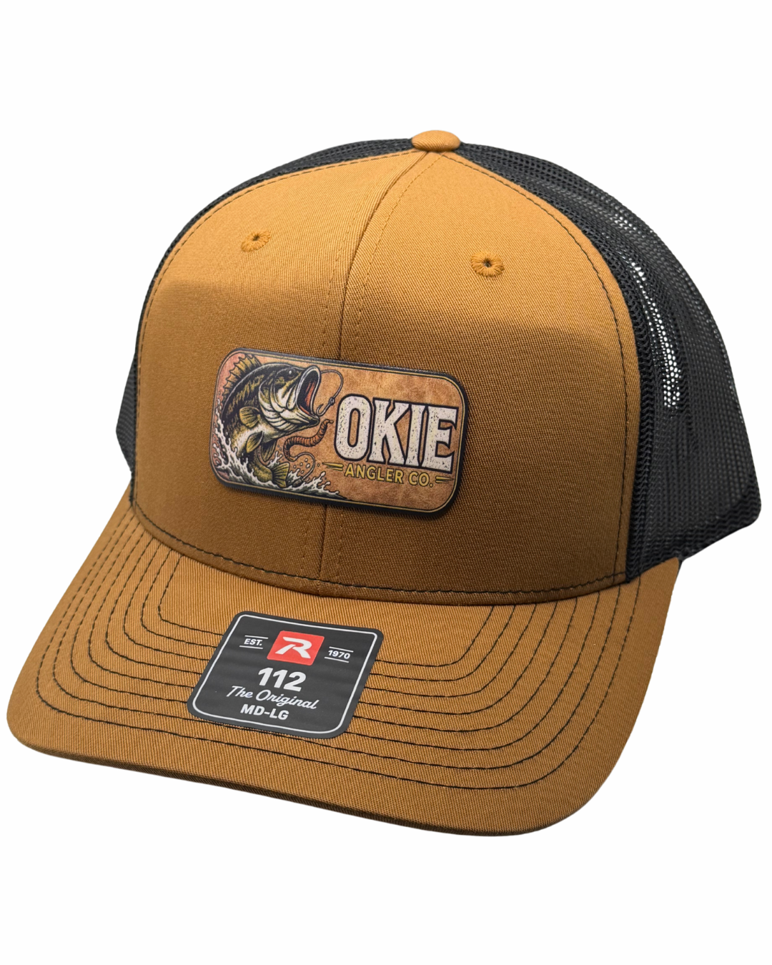 A trucker-style baseball cap with a mustard yellow front and brim, black mesh back panels, and a rectangular embroidered patch on the front that reads 'OKIE MADE' with a fish illustration. A size/brand sticker is visible on the brim.