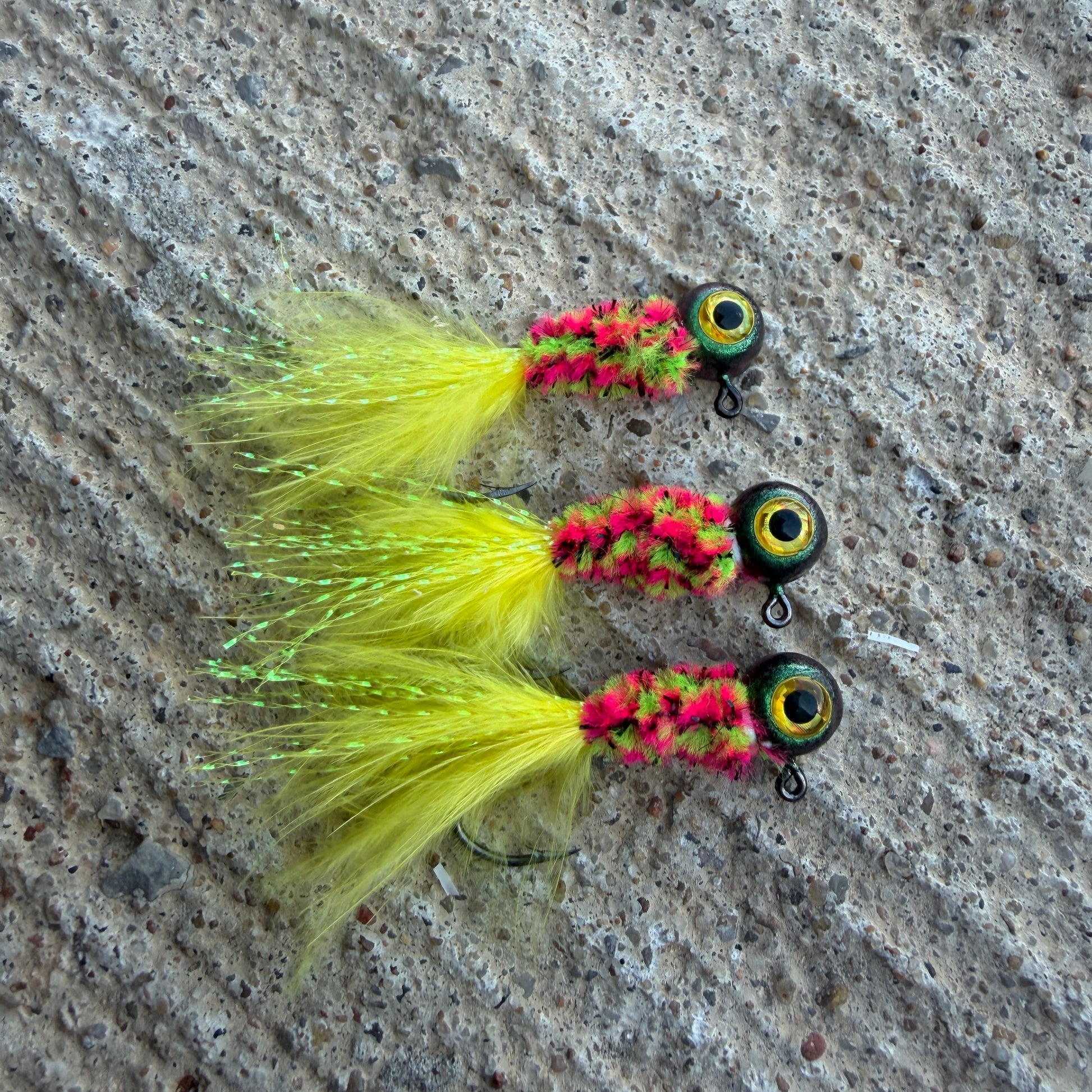 Three crappie fishing jigs with yellow and pink details on a textured surface.