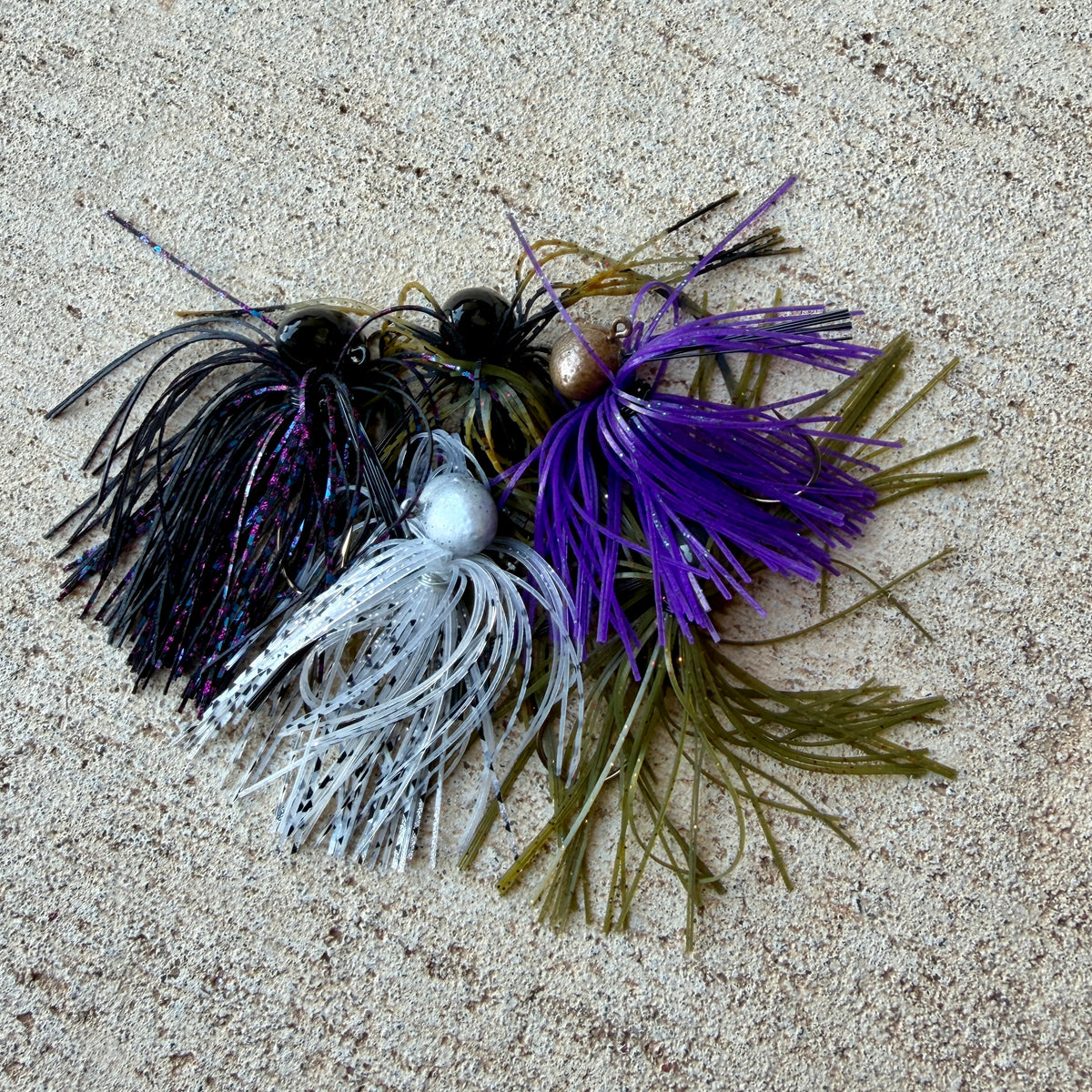 Four fishing jig lures with skirted heads and soft tails, arranged on a sandy beige surface. The lures include black and purple, white and silver, and olive green and purple combinations.