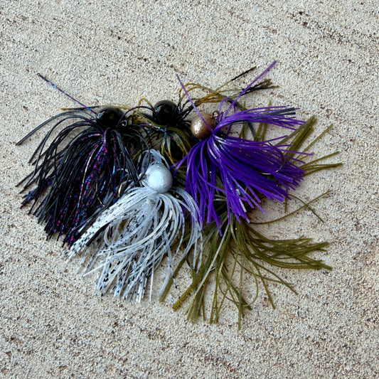 Four fishing jig lures with skirted heads and soft tails, arranged on a sandy beige surface. The lures include black and purple, white and silver, and olive green and purple combinations.