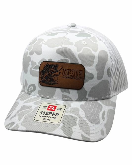 A white and gray camouflage trucker hat with a brown leather patch on the front featuring a fish graphic and the text 'OKIE'. The back of the cap is made of white mesh, and a white sewn-in sticker on the brim reads '112PFP PRINTED PANEL OSFM'.